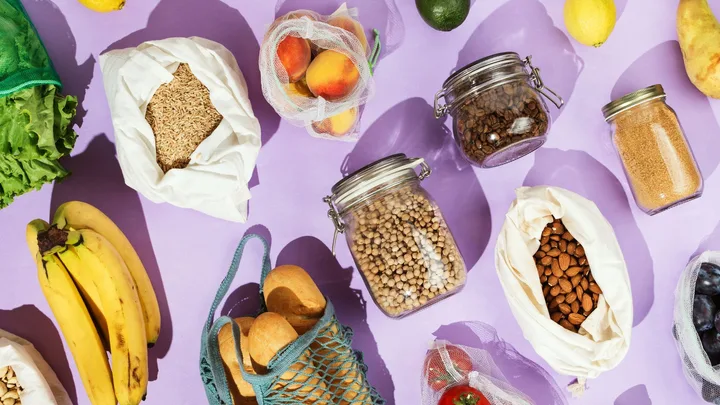 Assorted groceries on a purple background, including bananas, lettuce, grains, almonds, chickpeas, and reusable bags.