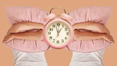 Person hugging pink pillow with alarm clock in center, on a beige background.