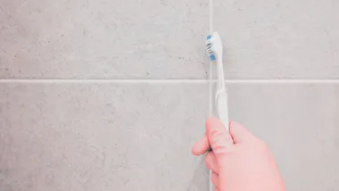 A hand in a pink glove using a toothbrush to clean grout between light-colored tiles.