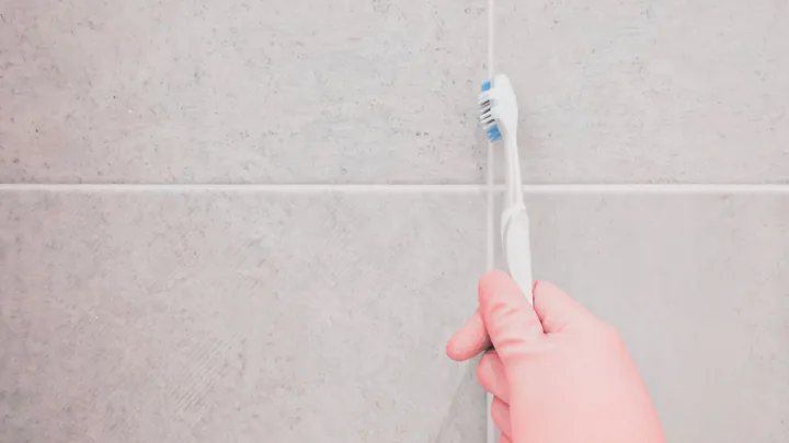 A hand in a pink glove using a toothbrush to clean grout between light-colored tiles.