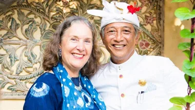 Two smiling people, a woman in a blue patterned scarf and a man in white attire with a floral headdress, stand together.