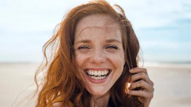 Smiling woman with red hair on a beach, sunlight on her face, looking directly at the camera.
