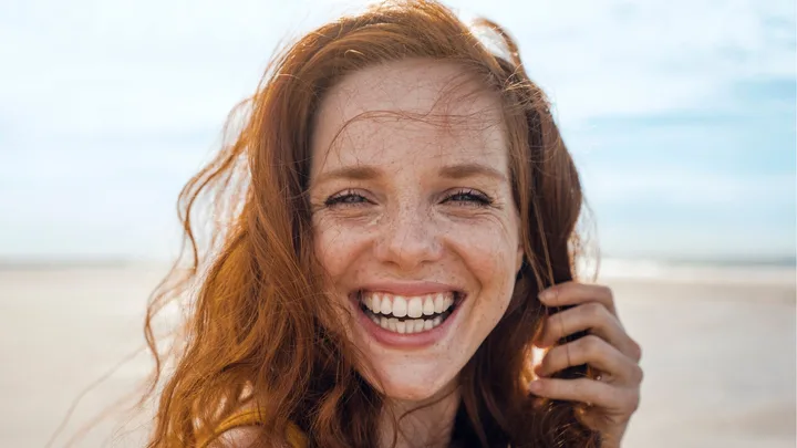 Smiling woman with red hair on a beach, sunlight on her face, looking directly at the camera.