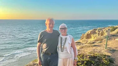Couple smiling by a seaside cliff at sunset with clear sky and ocean backdrop.