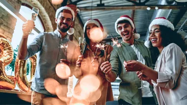 Group of four people in Santa hats celebrating with sparklers and champagne, against a festive background.