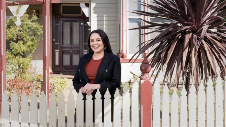 A woman in a black jacket smiles while standing behind a white picket fence in front of a house.
