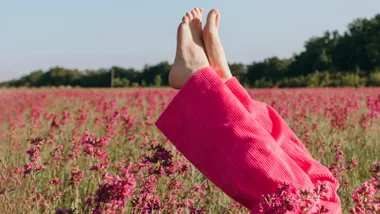 Bare feet in pink pants pointing skyward in a field of pink flowers against a clear sky and distant trees.