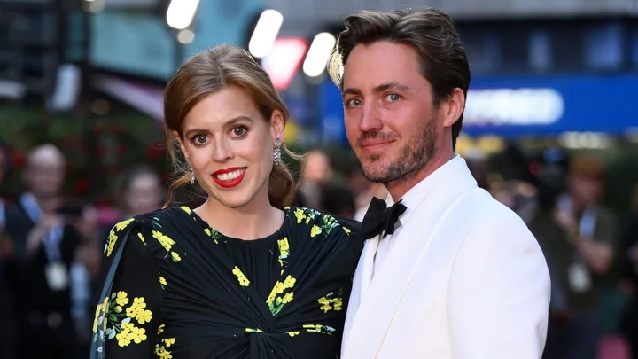 A woman in a floral dress and a man in a white tuxedo pose together on a red carpet.