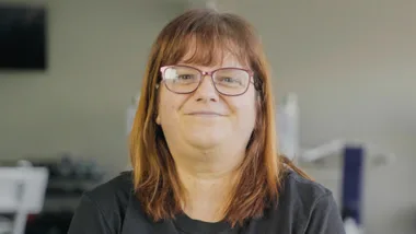 A woman with glasses and brown hair smiling at the camera indoors.