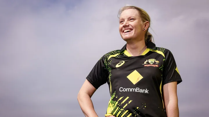 Woman in Australian cricket jersey smiling against a cloudy sky.