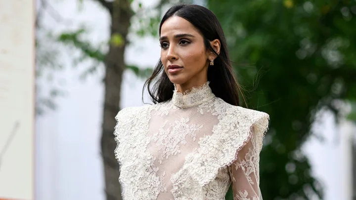 A woman in an elegant, lace-patterned outfit, standing outdoors with trees in the background.