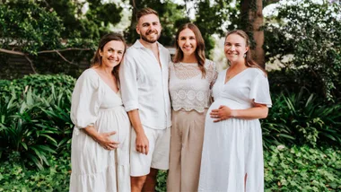 A group of four people, including two pregnant women, smiling in a garden setting with lush greenery.