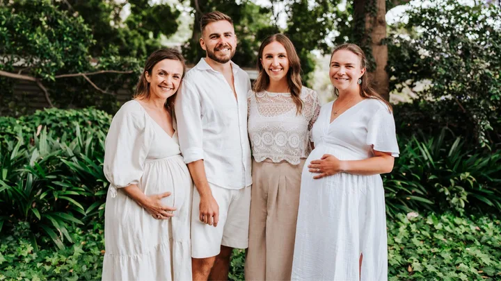 A group of four people, including two pregnant women, smiling in a garden setting with lush greenery.