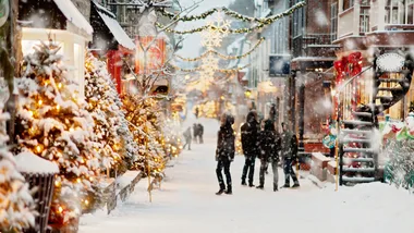 Snow-covered street with Christmas lights and decorations, people walking under snowflakes, festive trees lining the path.