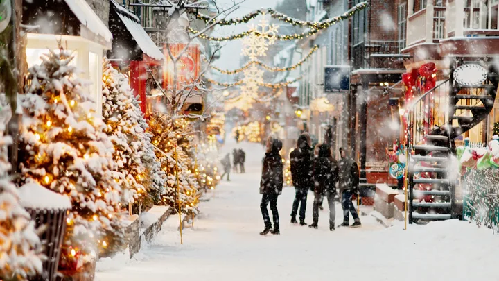 Snow-covered street with Christmas lights and decorations, people walking under snowflakes, festive trees lining the path.