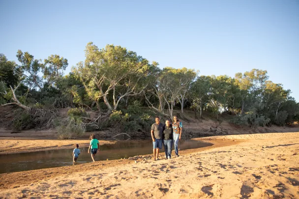 Three people stand on a sandy riverbank with trees in the background, while two others wade in shallow water.
