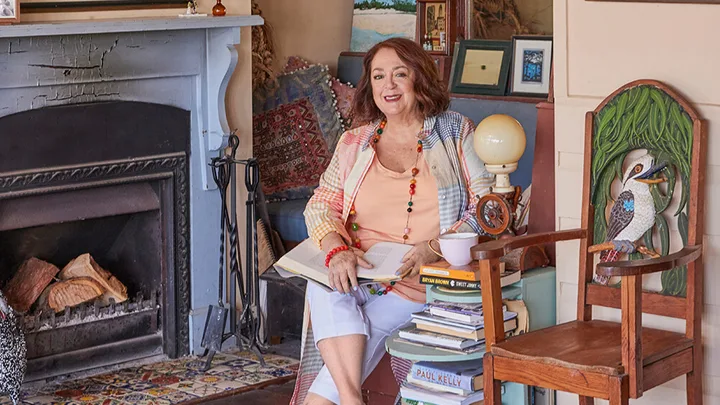 A woman sits by a fireplace with books around her, smiling warmly in a cozy room.