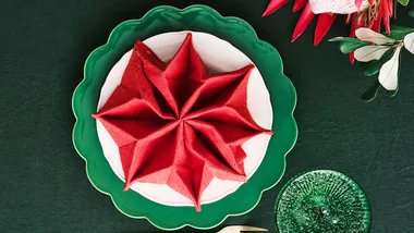 Red cloth napkin folded like a star on white plate with green trim, set on green tablecloth, with floral accents.