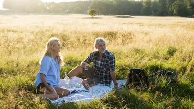 A couple enjoys a picnic on a blanket in a sunlit field, surrounded by tall grass and serene nature.