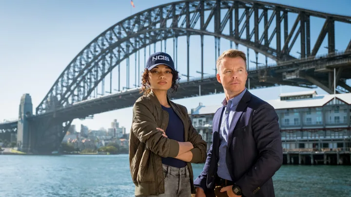 Two actors from "NCIS: Sydney" stand in front of the Sydney Harbour Bridge, one wearing an NCIS cap.