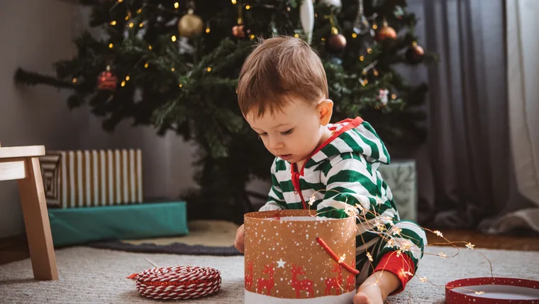 Toddler in striped pajamas explores a gift box near a festive Christmas tree decorated with lights and ornaments.