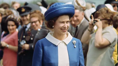 A woman in a blue outfit and hat smiles while walking outdoors, with a crowd in the background.
