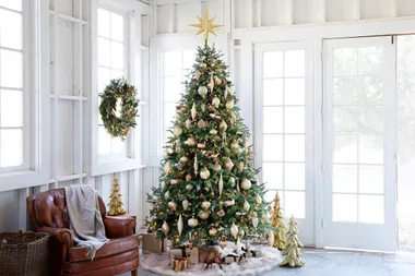 A decorated Christmas tree with gold ornaments, gifts, and a wreath on the wall in a bright room.