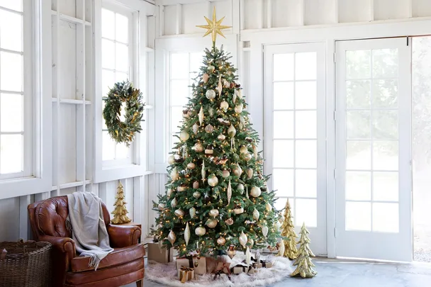 A decorated Christmas tree with gold ornaments, gifts, and a wreath on the wall in a bright room.