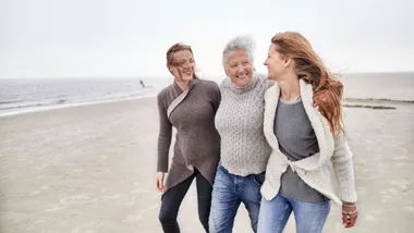 Three women walking and smiling on a windy beach, wearing cozy sweaters and jeans.