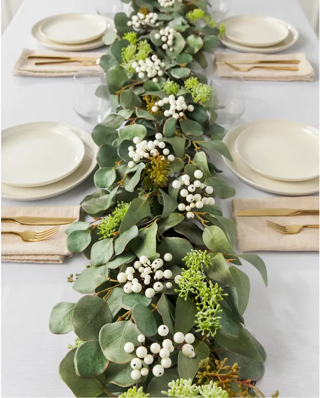 A table with a green eucalyptus and white berry centerpiece, set with white plates and gold cutlery on beige napkins.