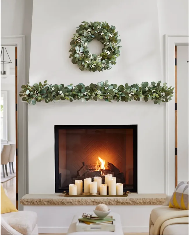 Fireplace with burning fire, decorated with eucalyptus wreath and garland, surrounded by candles on the mantel.