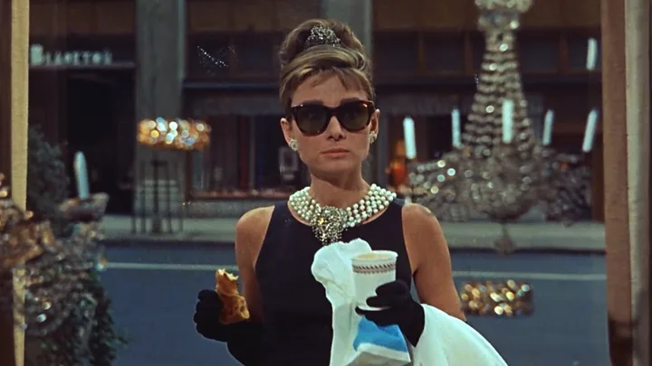 Elegant woman in black dress wearing sunglasses and pearls, holding coffee and pastry in city setting.