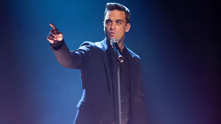 Singer performing on stage, holding a microphone and pointing dramatically with a blue spotlight in the background.