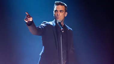 Singer performing on stage, holding a microphone and pointing dramatically with a blue spotlight in the background.