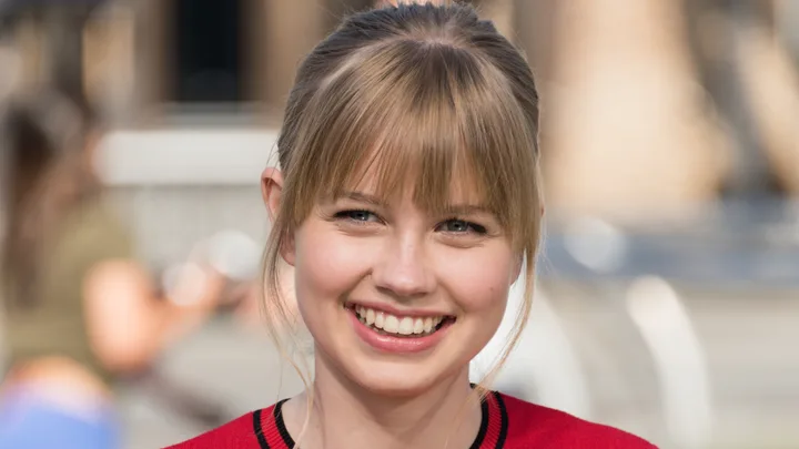 A smiling woman with blonde hair and bangs wears a red top outdoors.