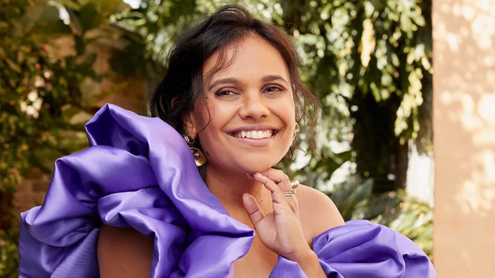 Smiling woman in a vibrant purple off-the-shoulder dress with ruffled sleeves, outdoors with green foliage in background.