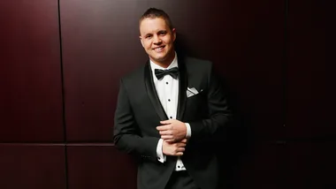 Man in a formal black tuxedo with a white shirt, standing against a dark wood-paneled wall, smiling.