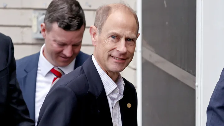 Man in suit smiles while walking outdoors, accompanied by another man in suit behind him.