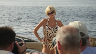 Woman in leopard print swimsuit poses on a boat with photographers taking pictures, set against an ocean backdrop.