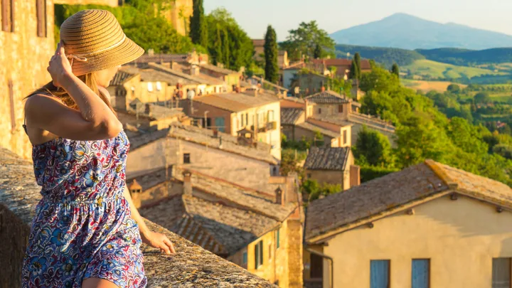 Woman in a floral dress and hat overlooking a picturesque hillside village with lush greenery and distant mountains.