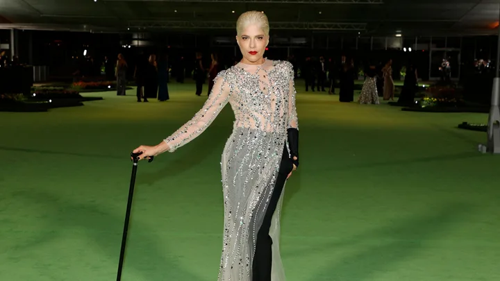 A person in an elaborate silver dress with red lipstick and a black cane, standing on a green carpet at a formal event.