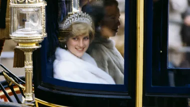 A woman in a tiara and white fur coat smiles inside a carriage, another woman sits beside her.