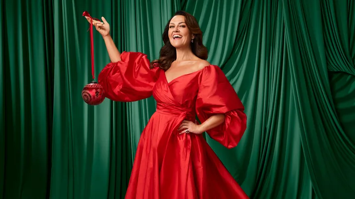A woman in a vibrant red dress holds a red ornament, standing against a green curtain background, smiling.