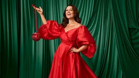 A woman in a vibrant red dress holds a red ornament, standing against a green curtain background, smiling.