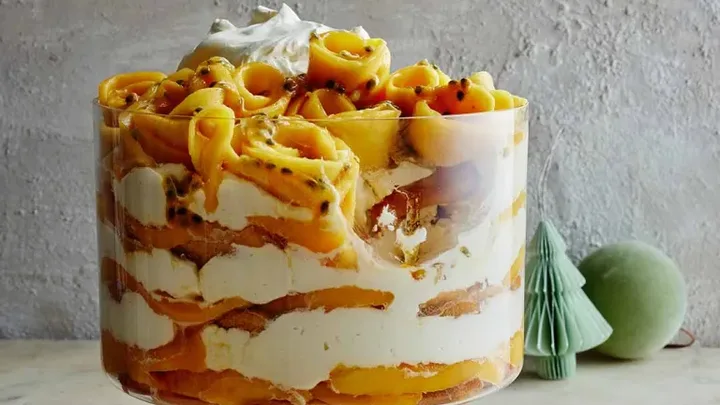 A layered tropical trifle with mango, passionfruit, and cream in a glass dish, with a gold spoon beside it on a table.