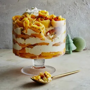 A layered tropical trifle with mango, passionfruit, and cream in a glass dish, with a gold spoon beside it on a table.