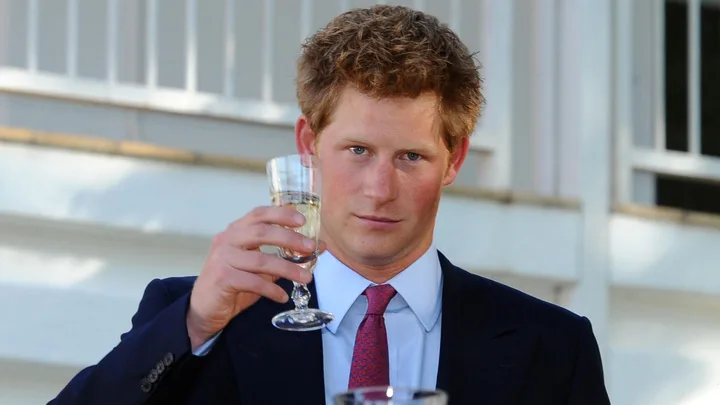 Man in a suit raising a glass in a toast, standing outdoors in front of a building with white siding.