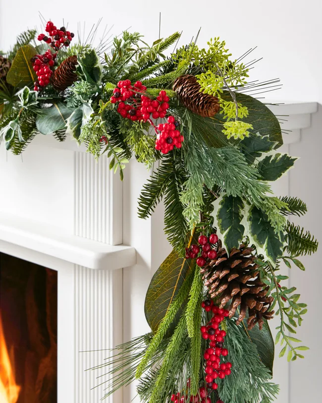 Festive holiday garland on a white mantel with pinecones, red berries, and greenery above a lit fireplace.