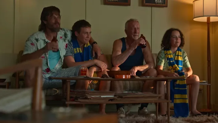 Four people sit on a couch wearing football scarves, watching TV, and holding drinks.