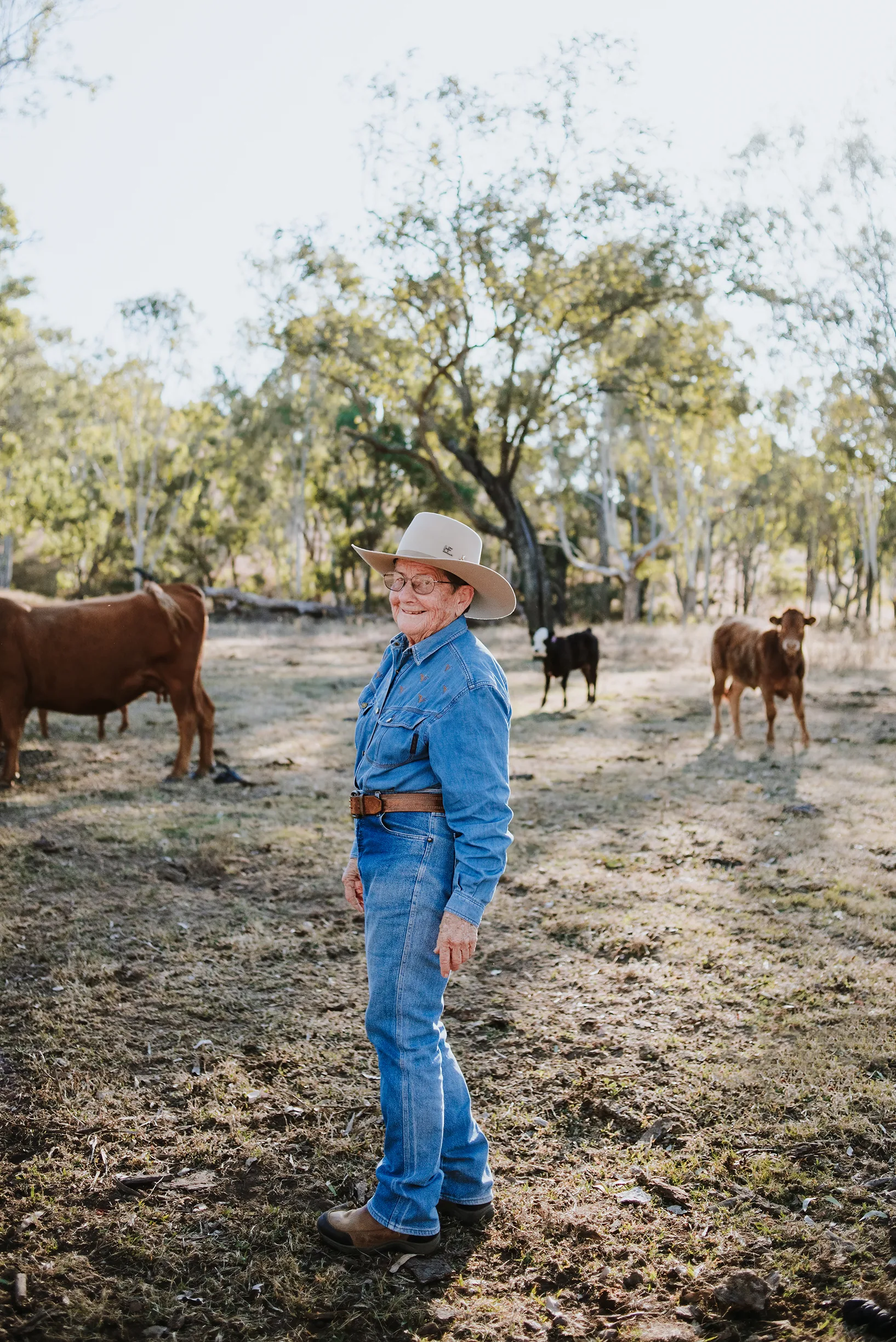 Girlie Goody (wearing denim jeans, a blue denim shirt and an Akubra hat) is standing in the back paddock with a herd of cattle.